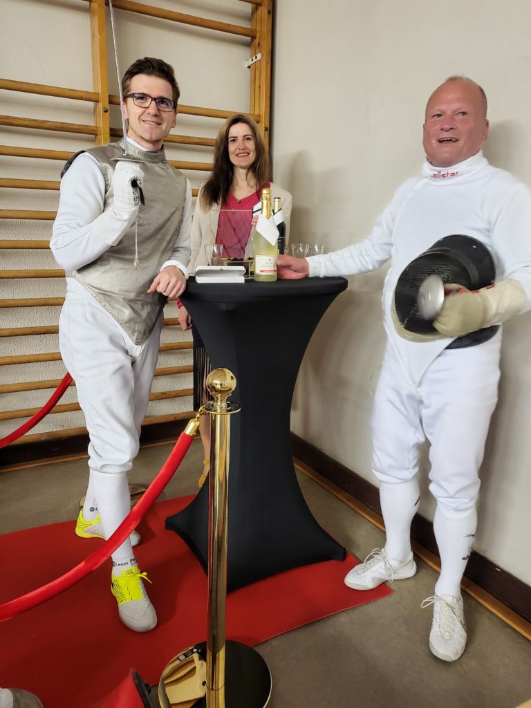 LUGART Arthur Schuchmilski und Anna Pianka von Designsie gratulieren dem Deutschen Meister in Florett 2022 in der Altersklasse V70, Berthold Schaum, mit einer Sektflasche mit BottleTie zur Auszeichung